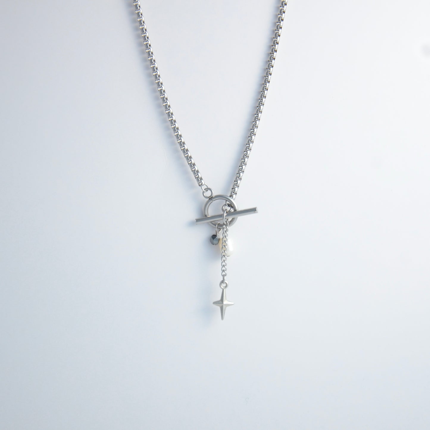Silver box chain necklace with a toggle clasp and a few charms including freshwater pearl, black glass bead and a dangling star chain on a light gray background