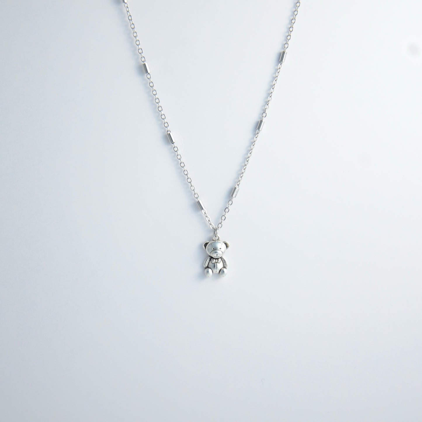 Dainty silver bar link chain with a teddy bear charm for minimalistic fashion or for layering jewelry on a light gray background
