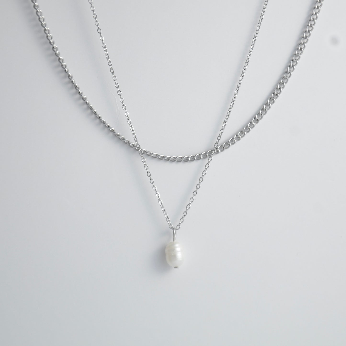 Silver stainless steel layered necklace set with a curb chain and a dainty pearl pendant necklace on a light gray background