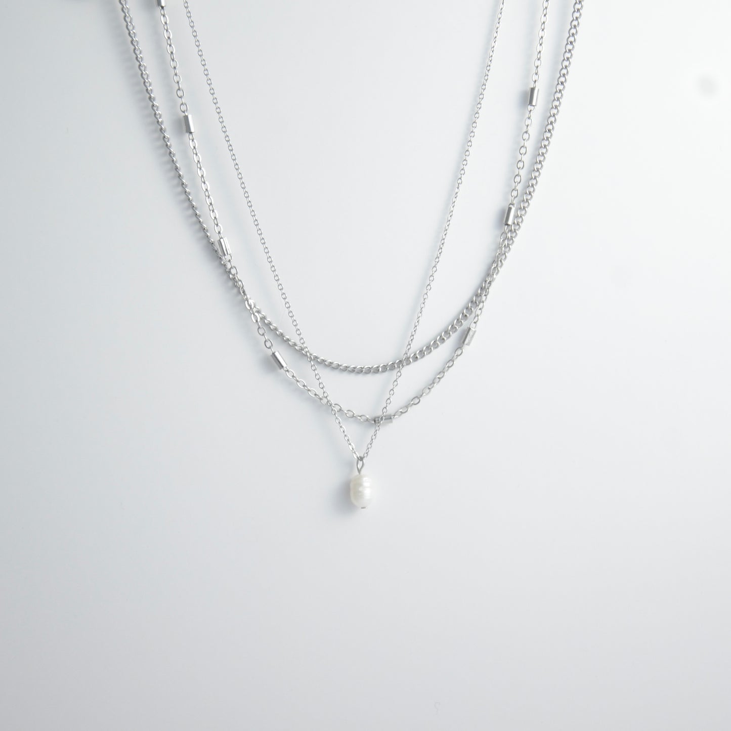 Silver stainless steel layered necklace set with a curb chain, a bar link chain and a dainty pearl pendant necklace on a light gray background