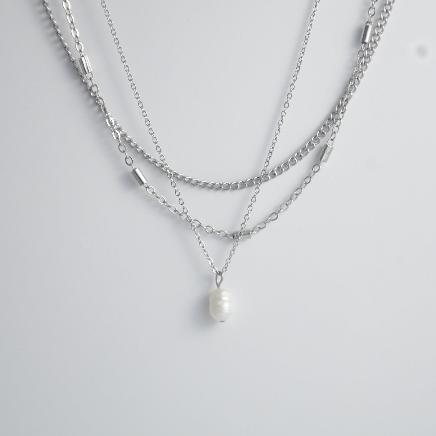 Silver stainless steel layered necklace set with a curb chain, a bar link chain and a dainty pearl pendant necklace on a light gray background