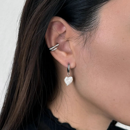 A close-up of a person's ear wearing a silver huggie hoop earring with a dangling white heart-shaped pearl, alongside a silver ear cuff.
