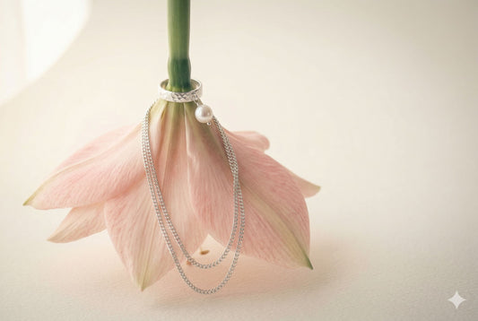 Sterling silver hammered ring with freshwater pearl charm and draped fine curb chains resting on blush pink amaryllis petals, spring jewelry editorial — Glass Balloon
