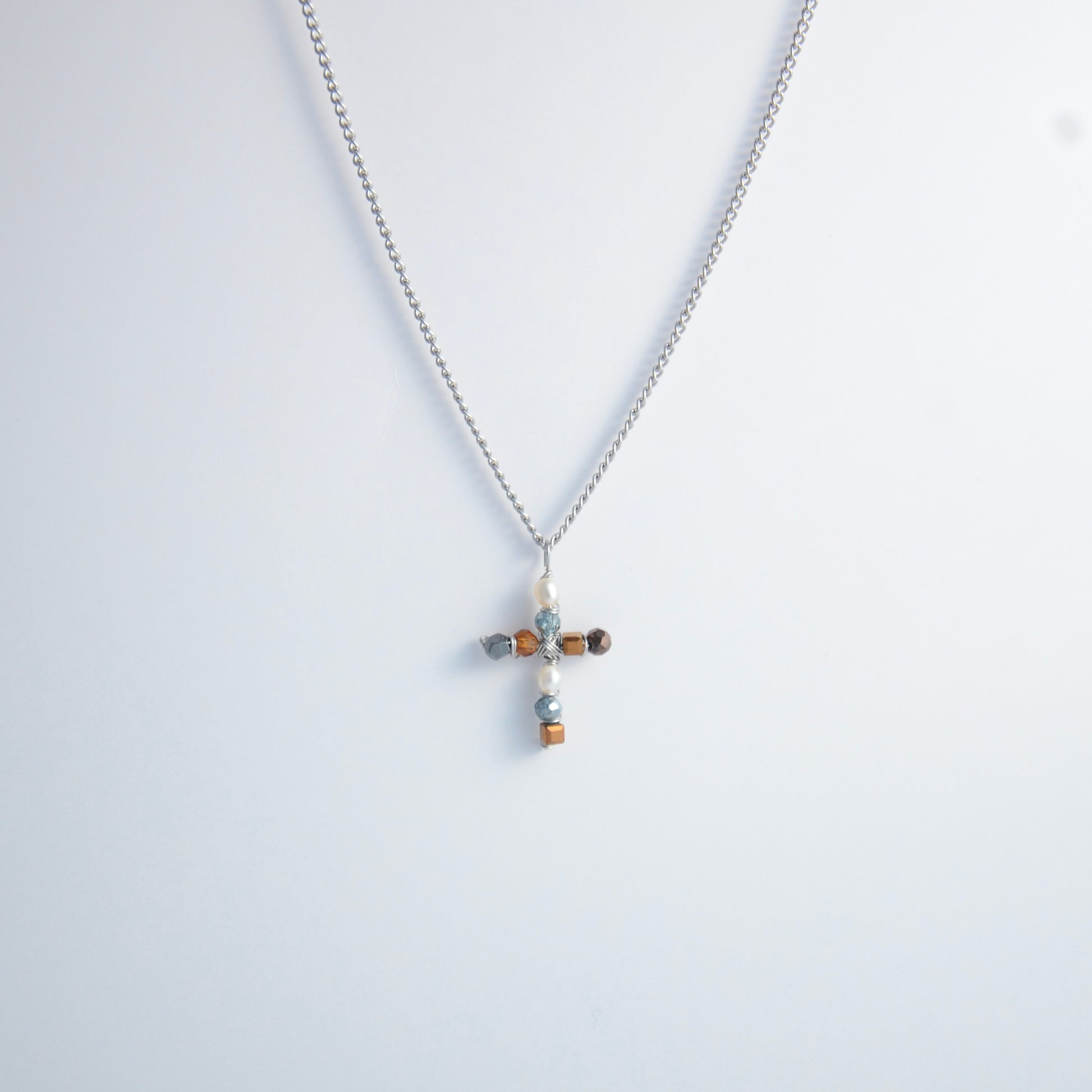 Silver curb chain necklace with a cross pendant made of freshwater pearls and blue, orange and black glass beads on a light gray background