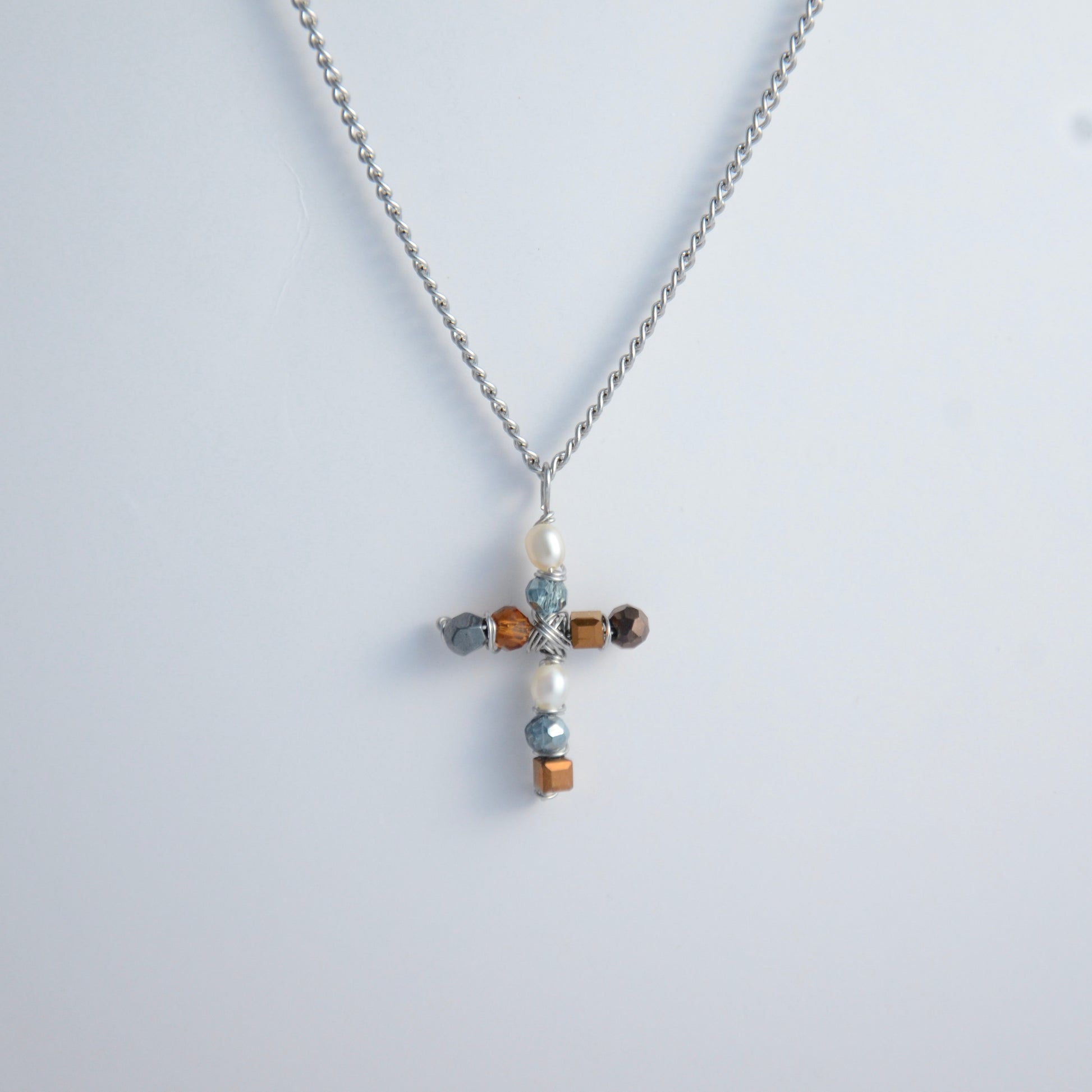 Silver curb chain necklace with a cross pendant made of freshwater pearls and blue, orange and black glass beads on a light gray background