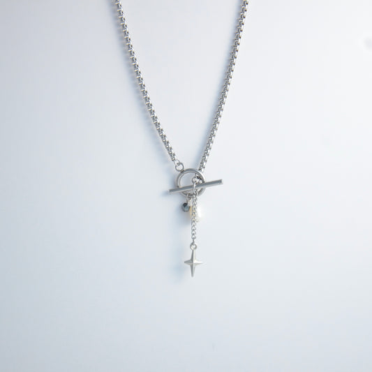 Silver box chain necklace with a toggle clasp and a few charms including freshwater pearl, black glass bead and a dangling star chain on a light gray background