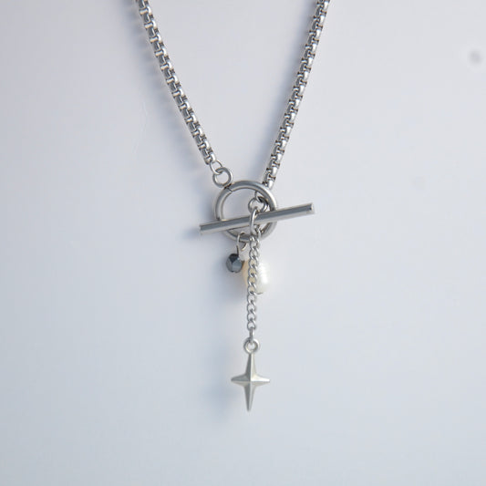 Silver box chain necklace with a toggle clasp and a few charms including freshwater pearl, black glass bead and a dangling star chain on a light gray background