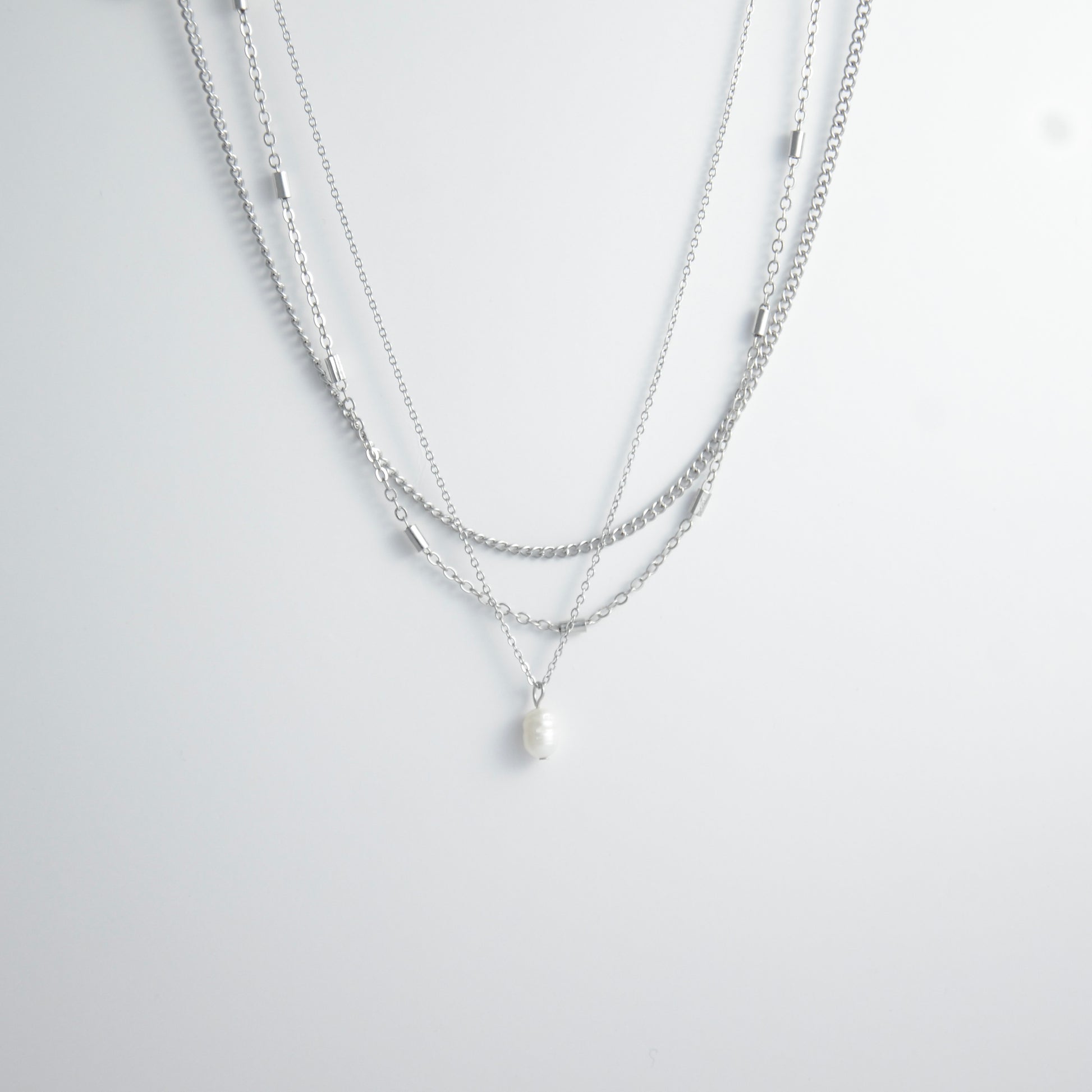 Silver stainless steel layered necklace set with a curb chain, a bar link chain and a dainty pearl pendant necklace on a light gray background