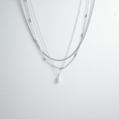 Silver stainless steel layered necklace set with a curb chain, a bar link chain and a dainty pearl pendant necklace on a light gray background