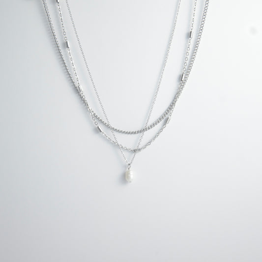 Silver stainless steel layered necklace set with a curb chain, a bar link chain and a dainty pearl pendant necklace on a light gray background