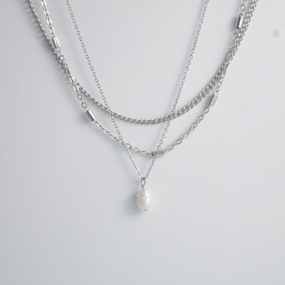 Silver stainless steel layered necklace set with a curb chain, a bar link chain and a dainty pearl pendant necklace on a light gray background