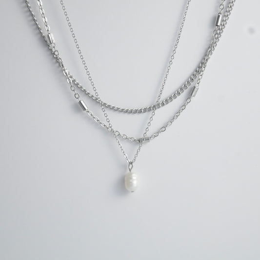 Silver stainless steel layered necklace set with a curb chain, a bar link chain and a dainty pearl pendant necklace on a light gray background