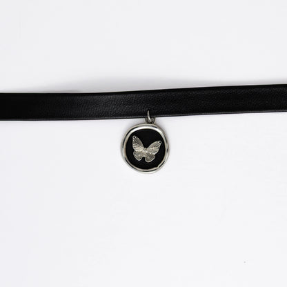 A black leather choker necklace with a central butterfly stamp pendant, displayed against a white background