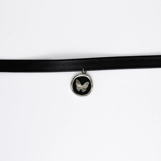 A black leather choker necklace with a central butterfly stamp pendant, displayed against a white background