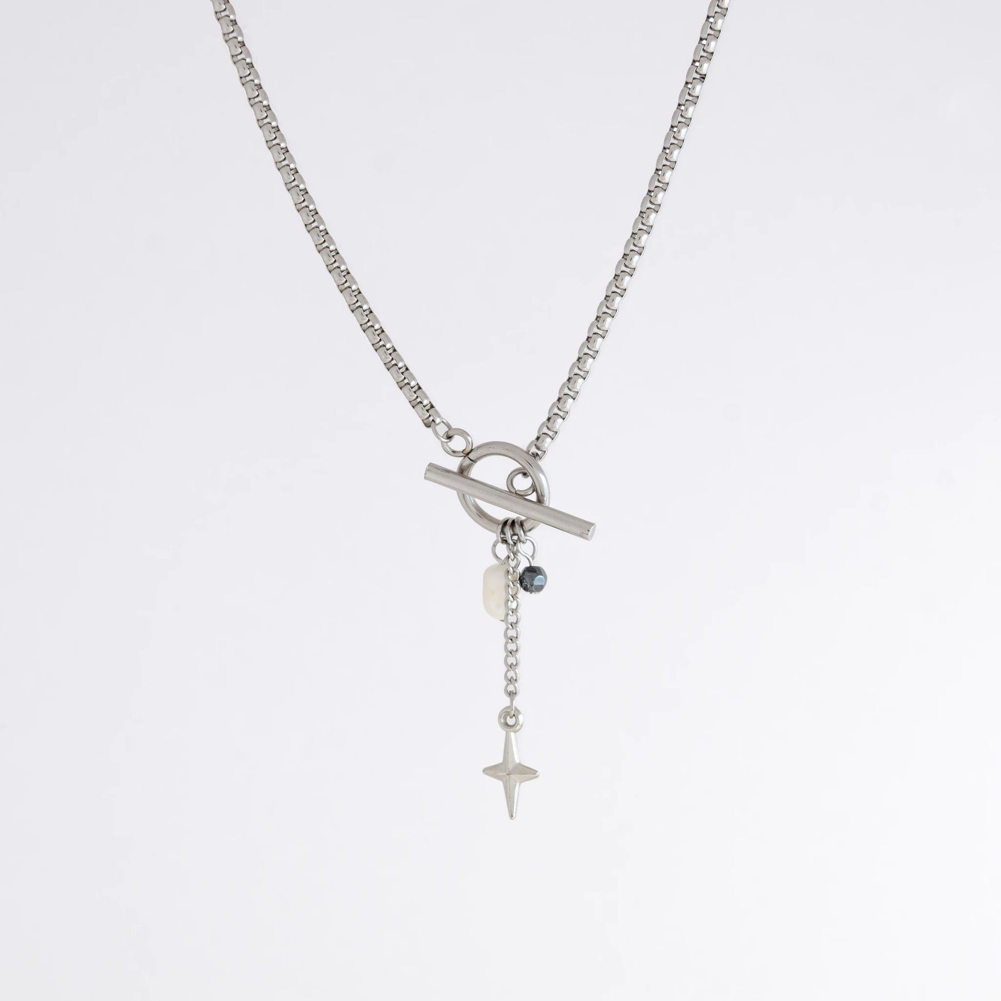 Silver box chain necklace with a toggle clasp and a few charms including freshwater pearl, black glass bead and a dangling star chain on a light gray background