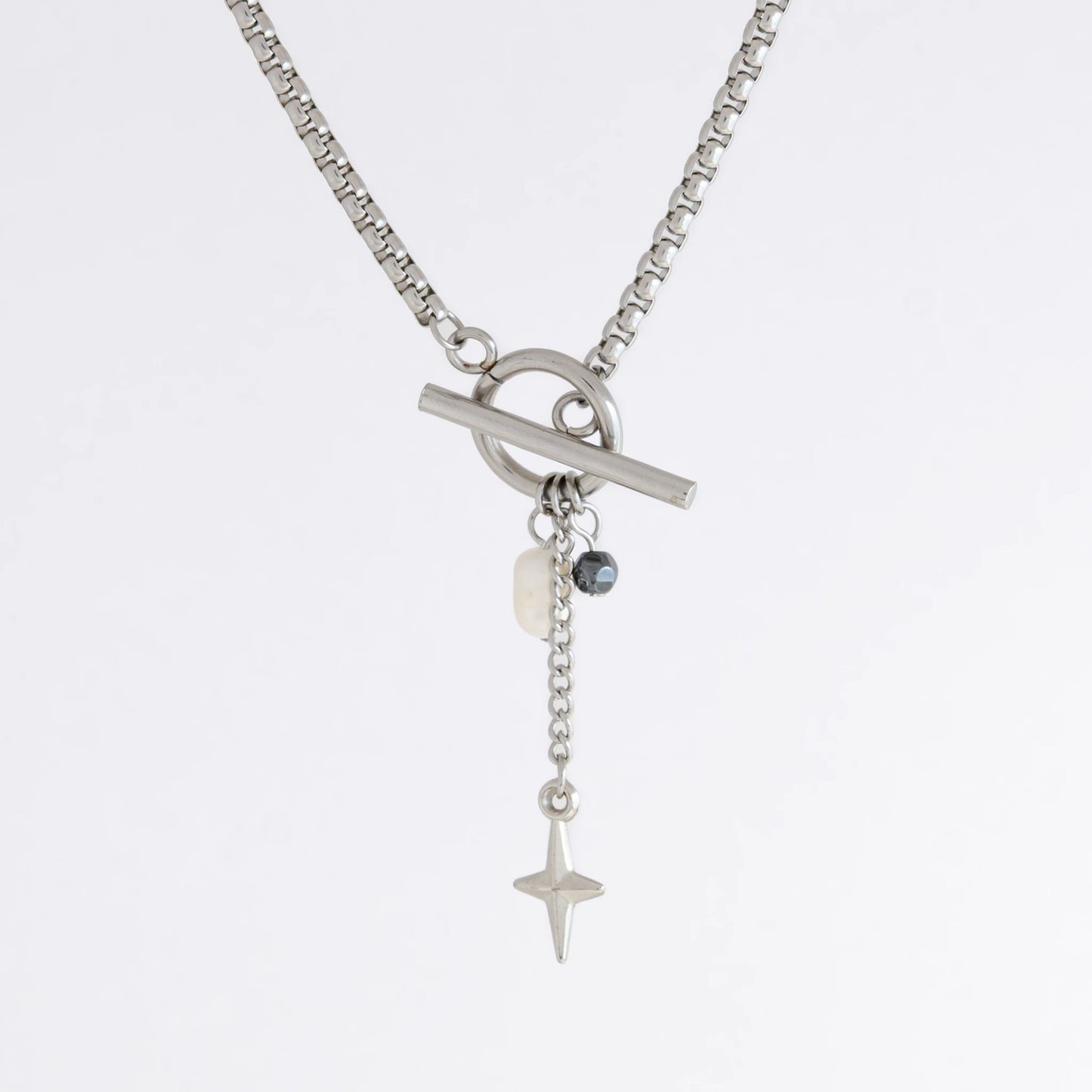 Silver box chain necklace with a toggle clasp and a few charms including freshwater pearl, black glass bead and a dangling star chain on a light gray background