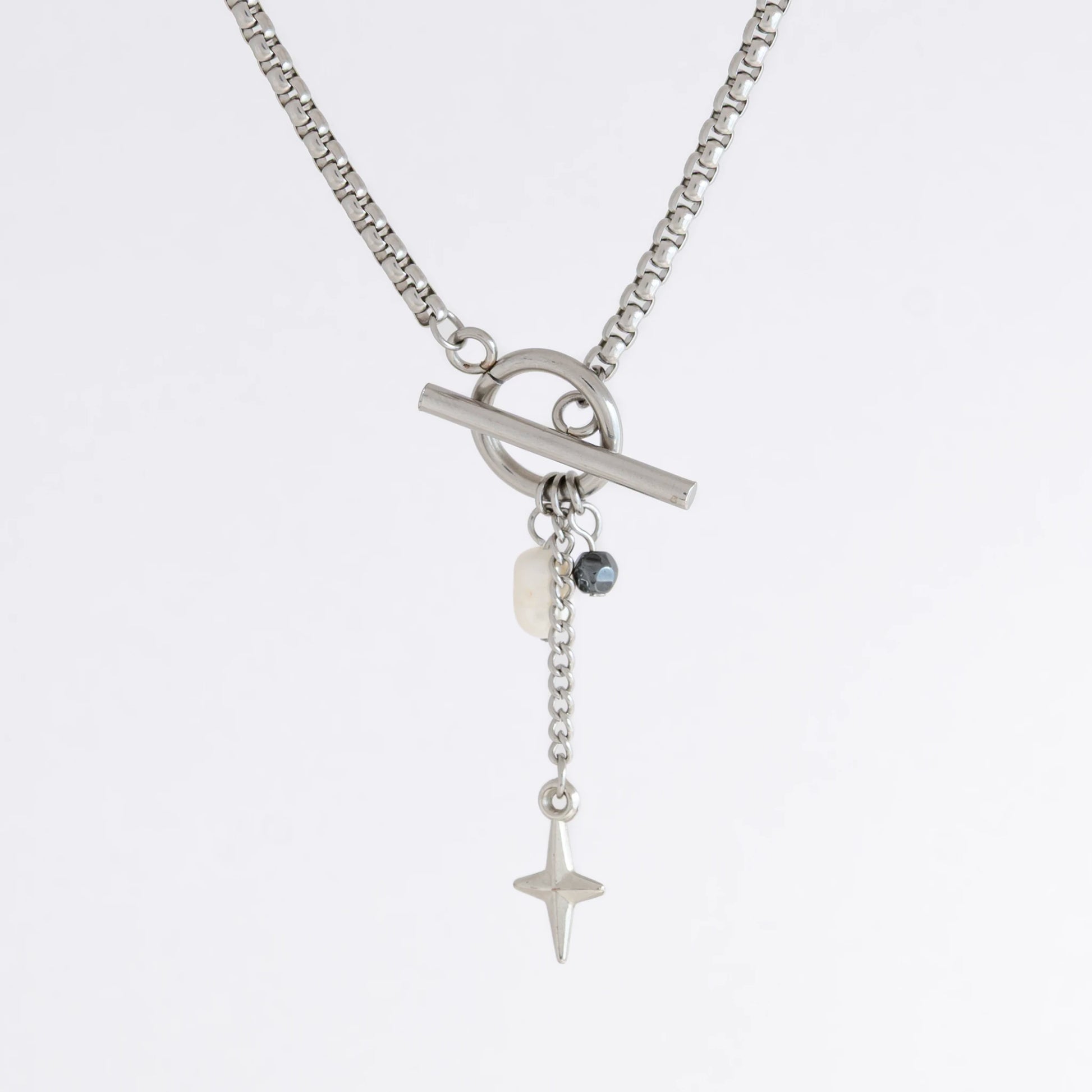 Silver box chain necklace with a toggle clasp and a few charms including freshwater pearl, black glass bead and a dangling star chain on a light gray background