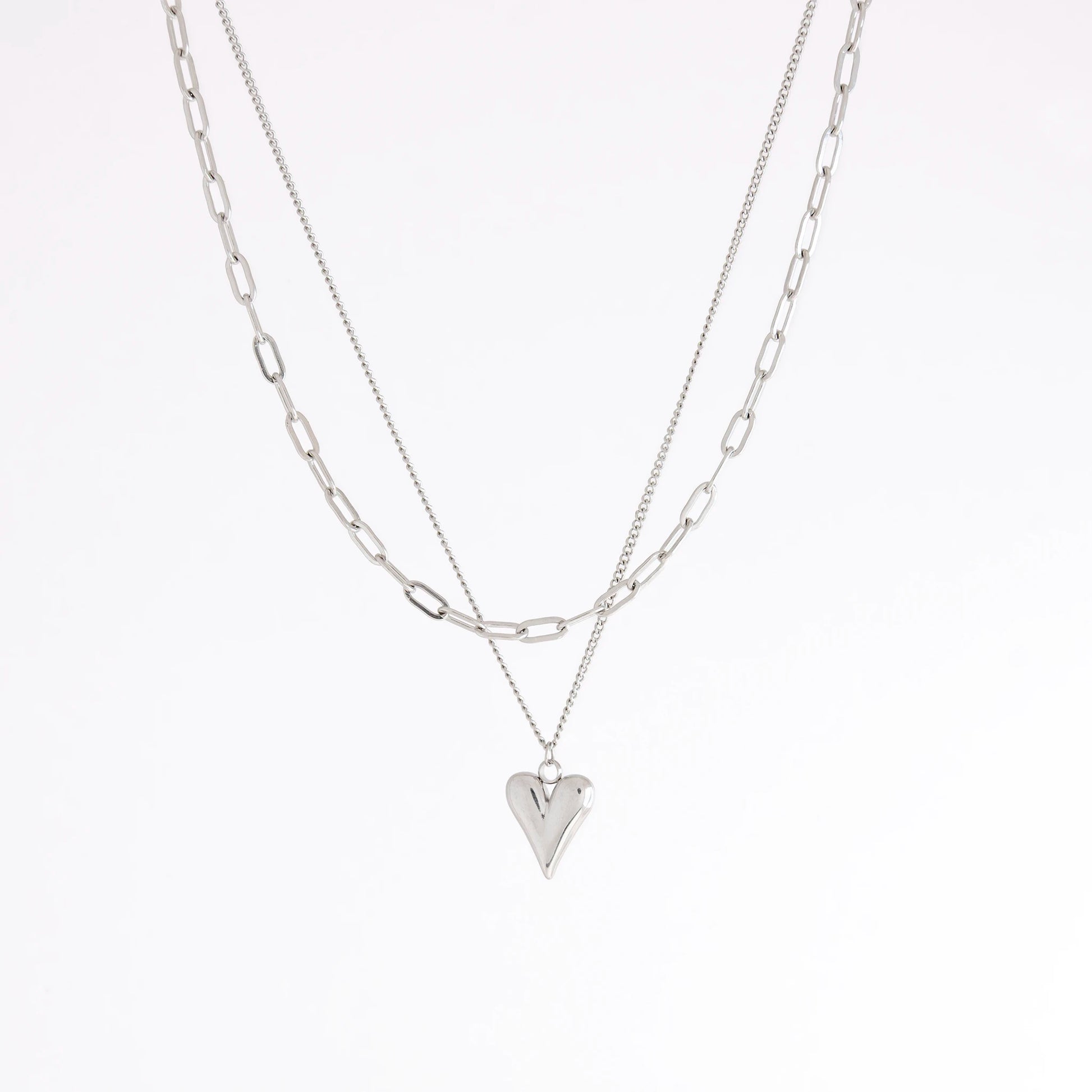 A layered silver necklace set featuring a paperclip chain and a chain with a small heart-shaped pendant, displayed against a clean white background.