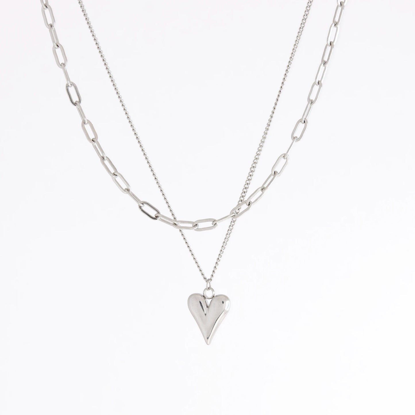 A layered silver necklace set featuring a paperclip chain and a delicate chain with a polished heart-shaped pendant, displayed on a clean white background.