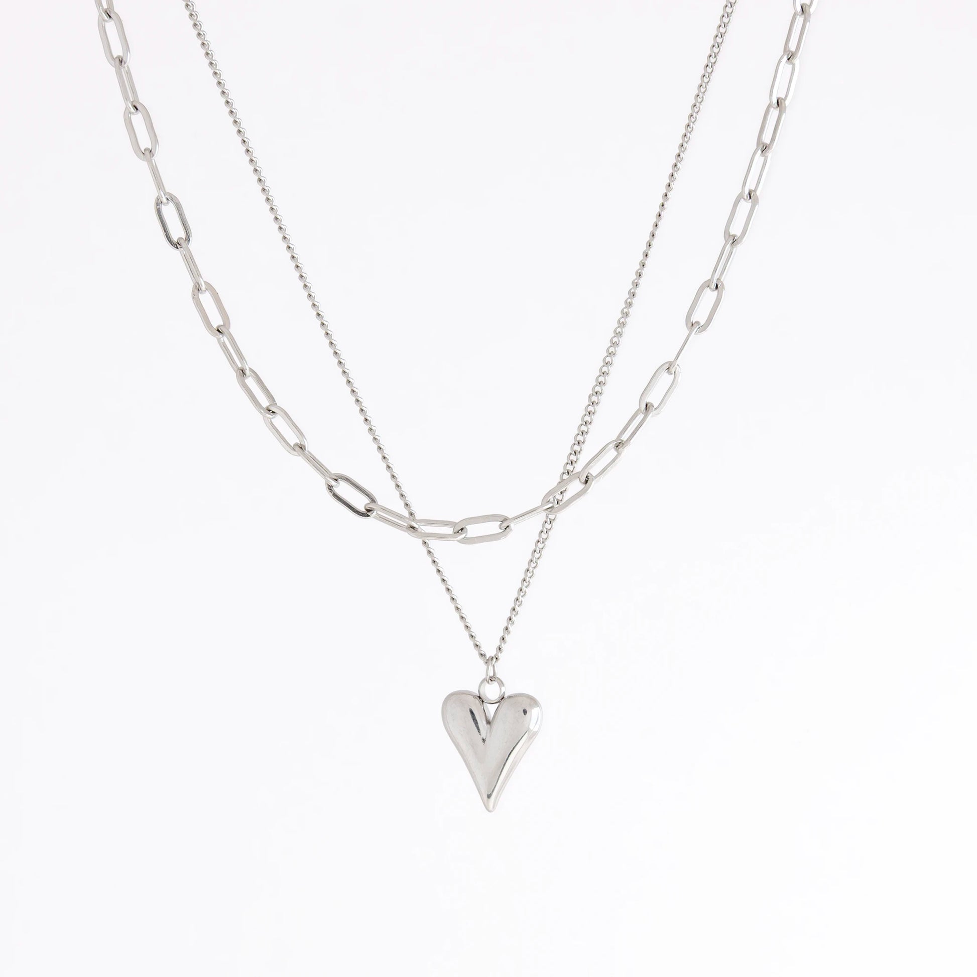 A layered silver necklace set featuring a paperclip chain and a delicate chain with a polished heart-shaped pendant, displayed on a clean white background.