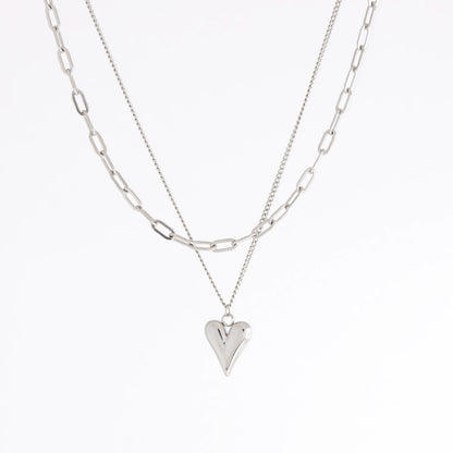 A layered silver necklace set featuring a paperclip chain and a delicate chain with a polished heart-shaped pendant, displayed on a clean white background.