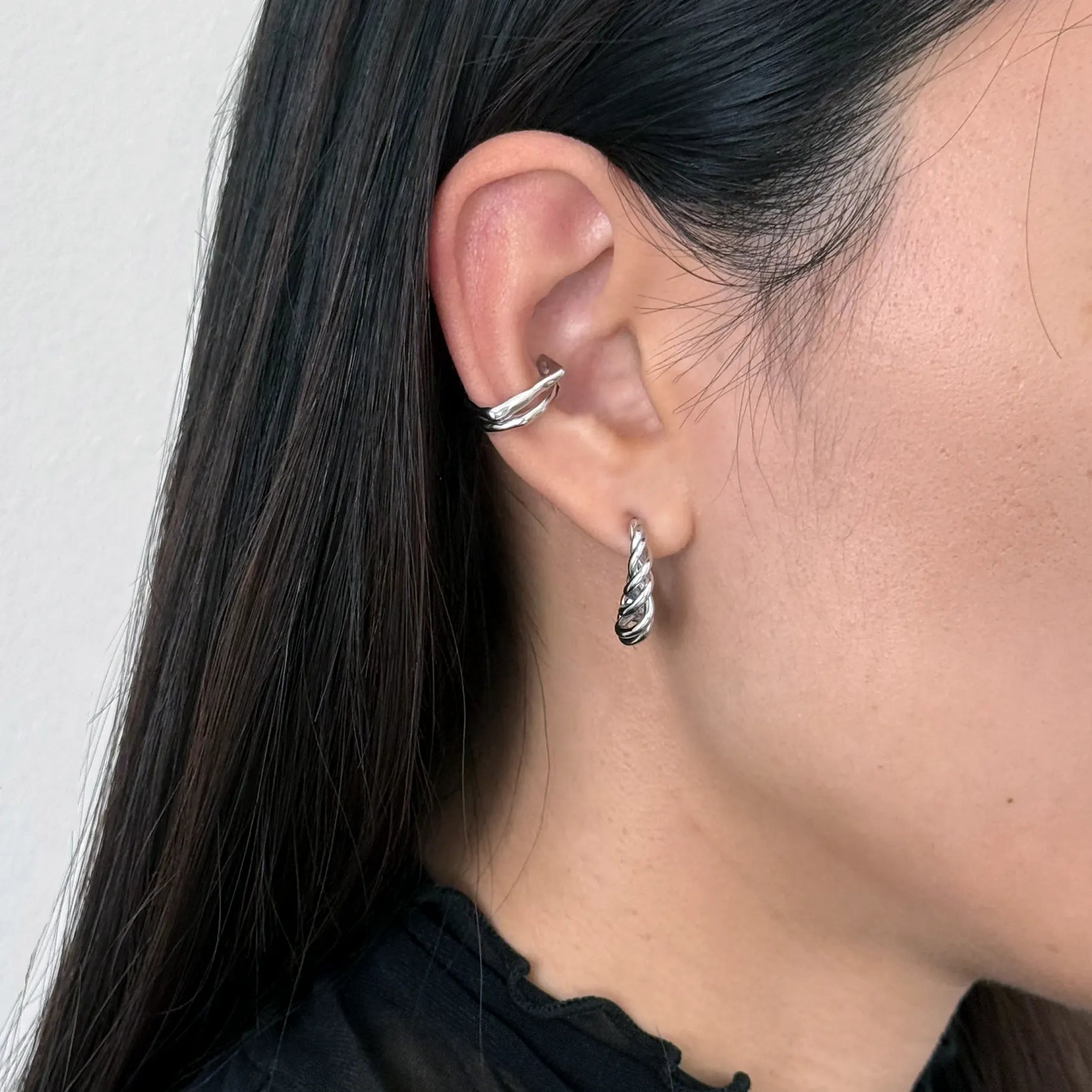 A close-up of a person's ear showcasing two silver earrings: a twisted huggie hoop in the lobe and a small twisted drop hoop on the lobe.