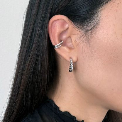 A close-up of a person's ear showcasing two silver earrings: a twisted huggie hoop in the lobe and a small twisted drop hoop on the lobe.
