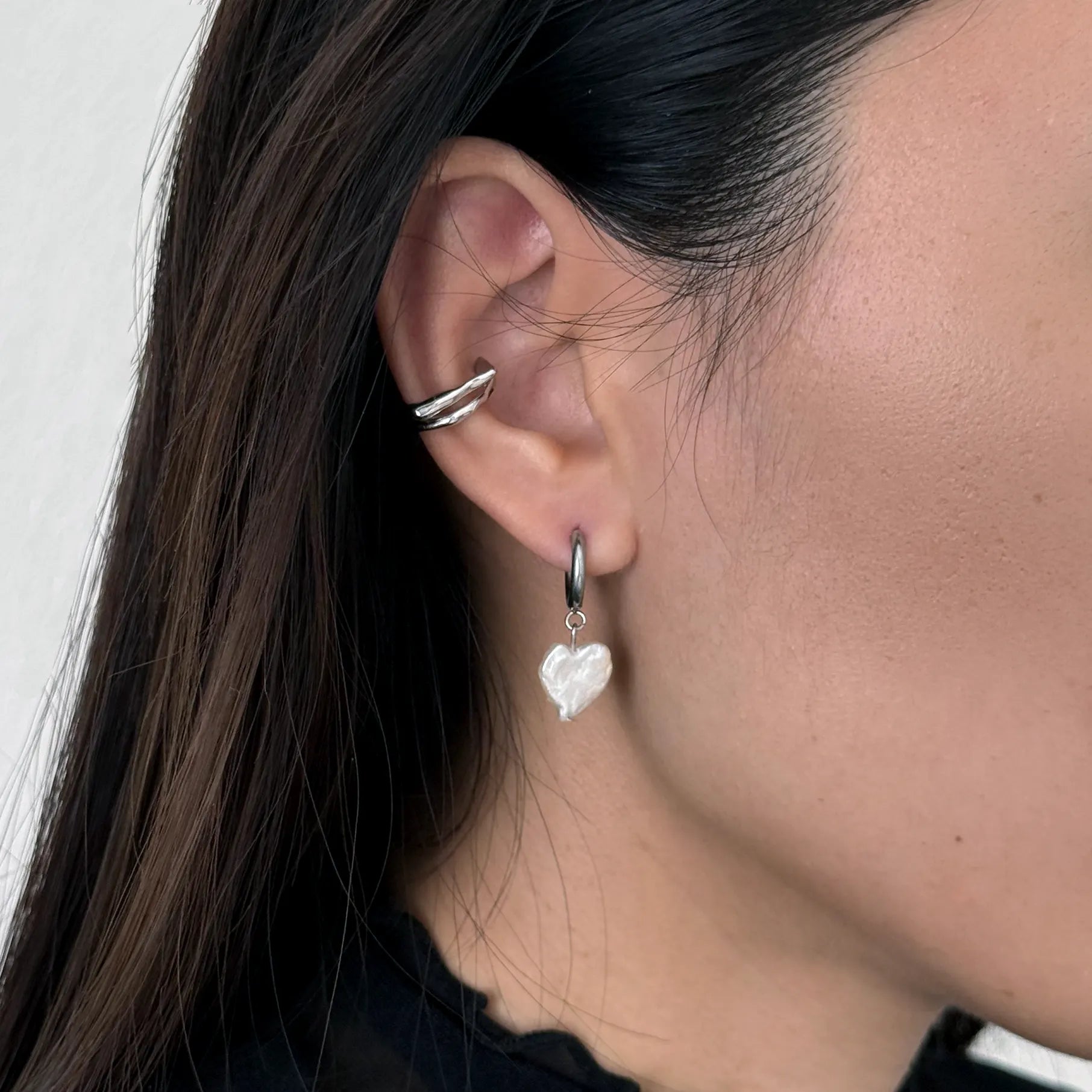 A close-up of a person's ear wearing a silver huggie hoop earring with a dangling white heart-shaped pearl, alongside a silver ear cuff.