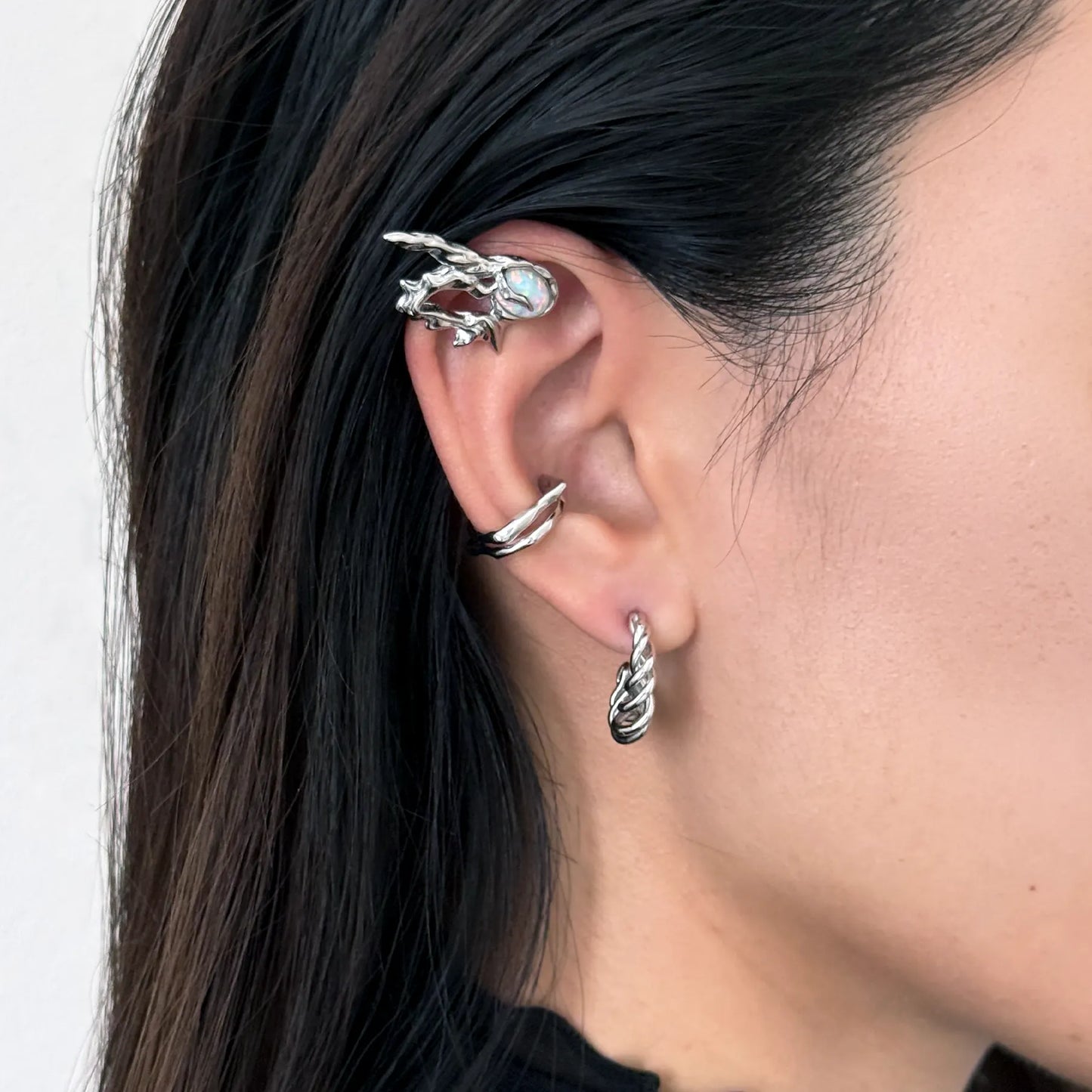 A close-up of a model's ear showcasing a stack of silver-toned ear cuffs, including a twisted spiral hoop in the lobe, a thin wrap-around cartilage cuff, and a larger claw-style ear cuff with a light blue stone, set against dark hair and a neutral background.