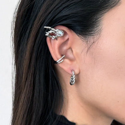 A close-up of a model's ear showcasing a stack of silver-toned ear cuffs, including a twisted spiral hoop in the lobe, a thin wrap-around cartilage cuff, and a larger claw-style ear cuff with a light blue stone, set against dark hair and a neutral background.