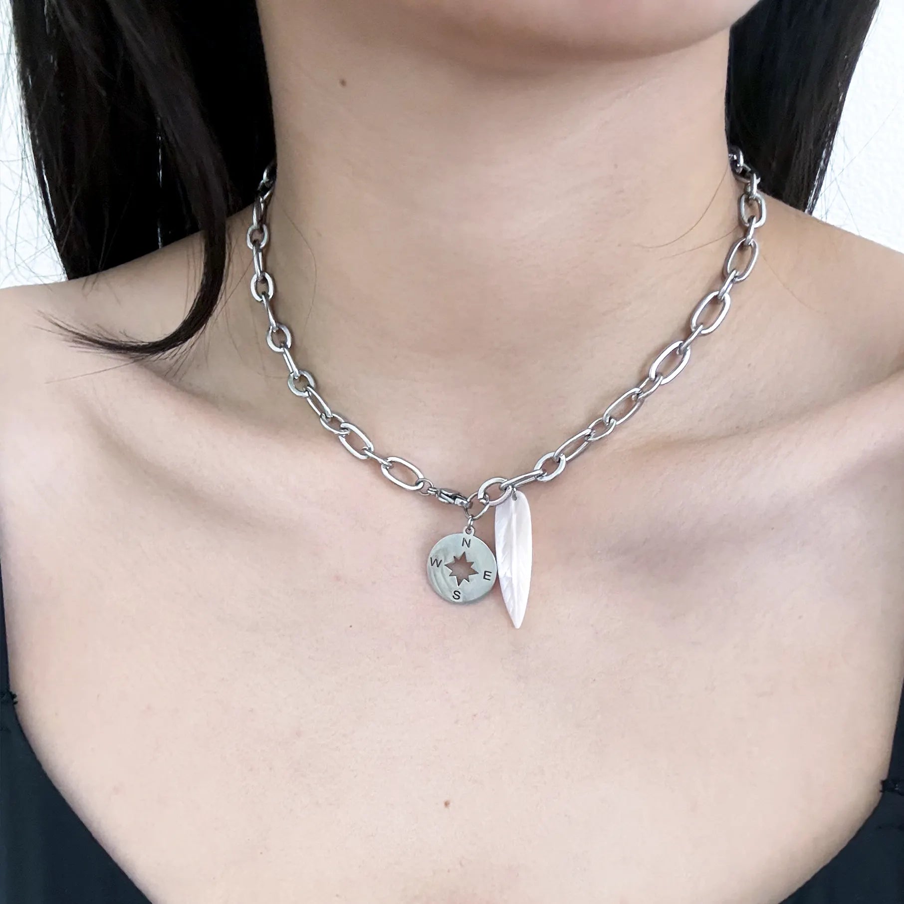 A close-up of a model wearing a silver chain necklace with a circular compass coin charm and a white teardrop crystal pendant.