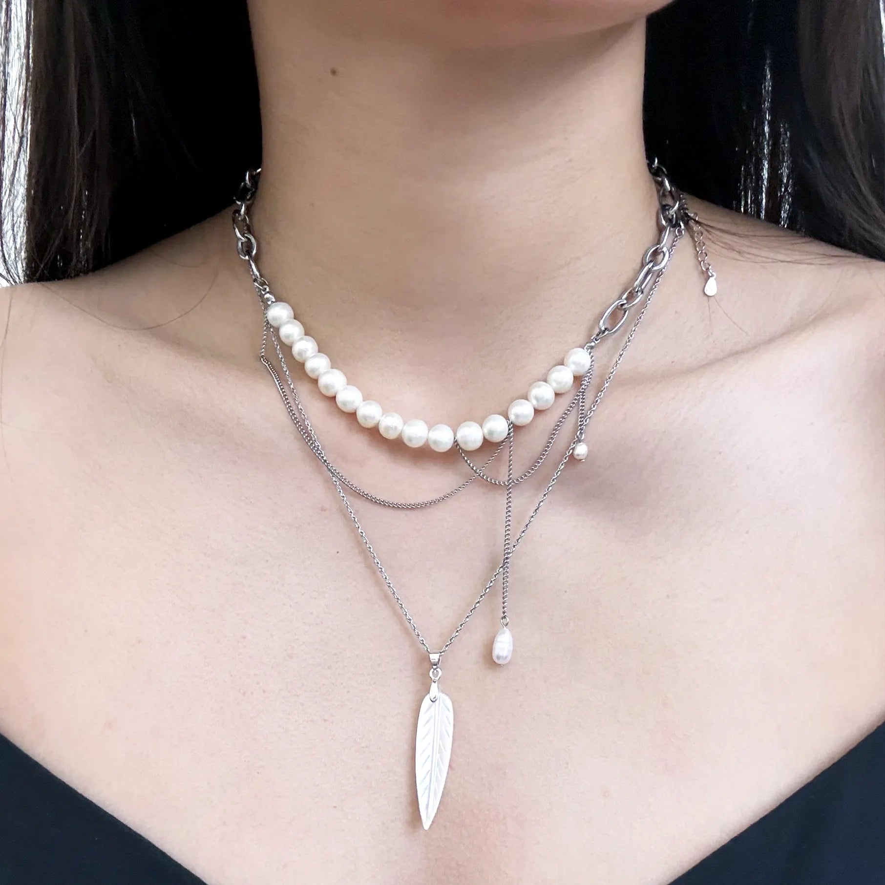 A close-up of a model wearing a multi-layered necklace set. The set includes a short pearl strand on a silver chain, a delicate silver chain with a silver feather pendant, and a thin silver chain with a small pearl drop.