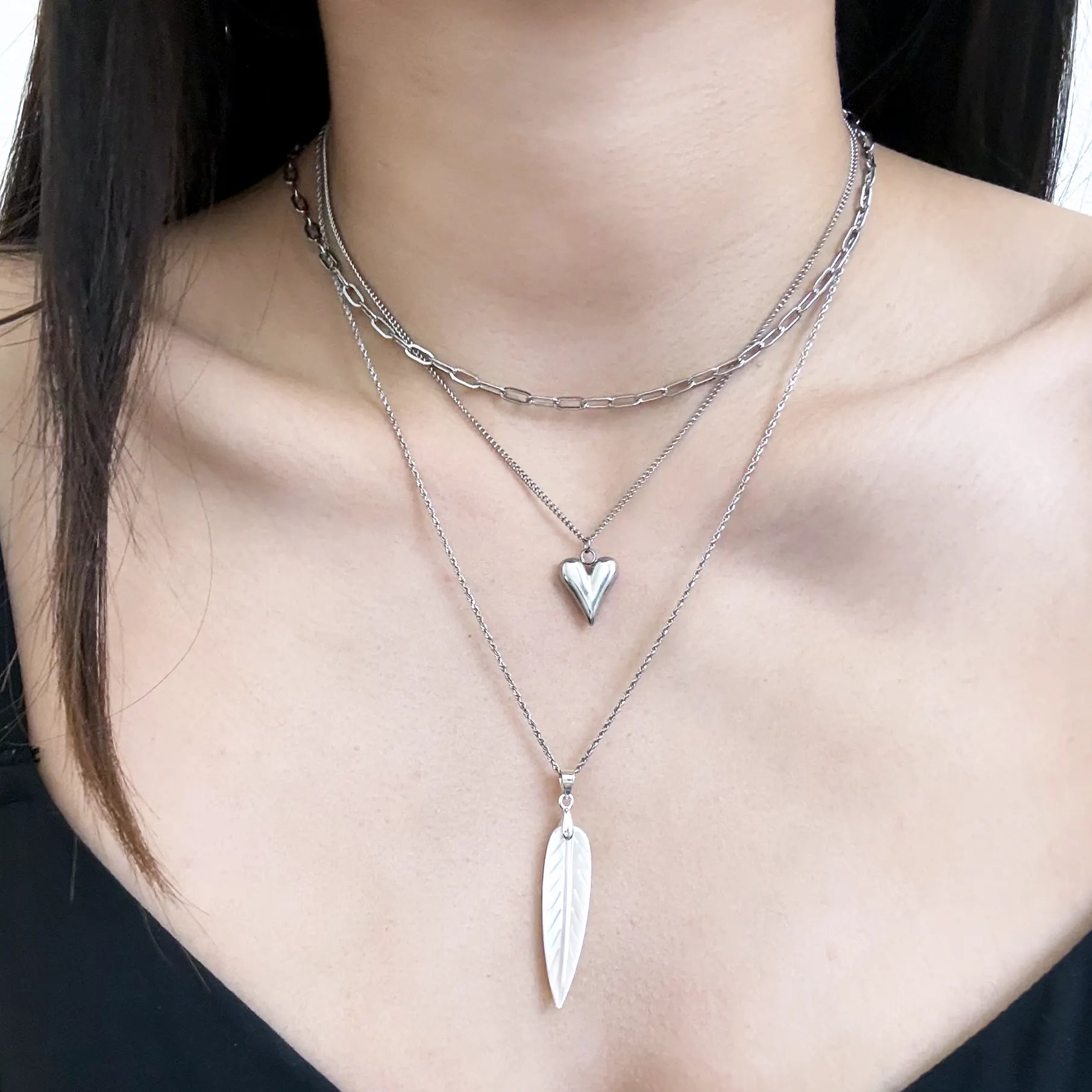A close-up of a person's neck and upper chest wearing three layered silver necklaces: a short curb chain, a paperclip chain, and a longer chain with a small heart pendant and a larger feather-shaped pendant.