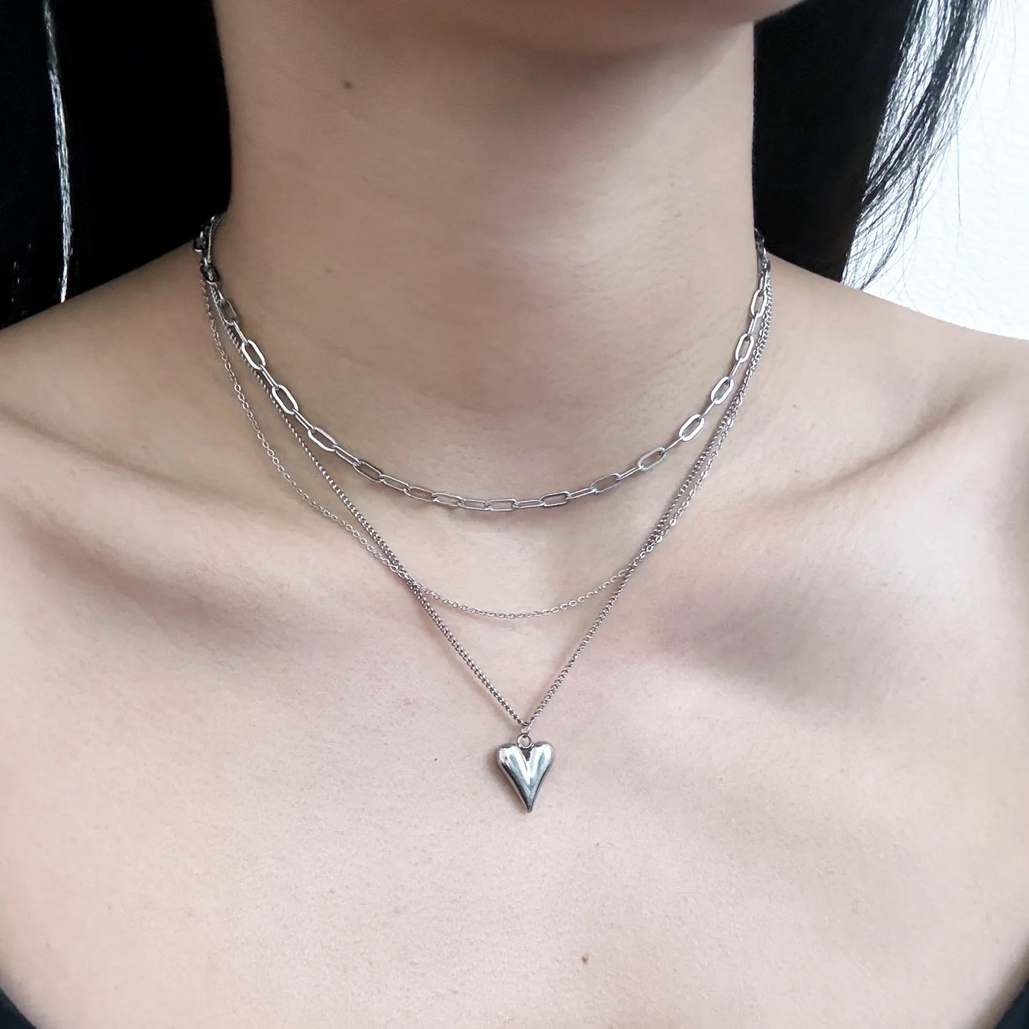 A close-up of a person's neck and upper chest wearing a layered silver necklace set, including a paperclip chain, a fine chain, and a pendant with a small heart-shaped cutout.