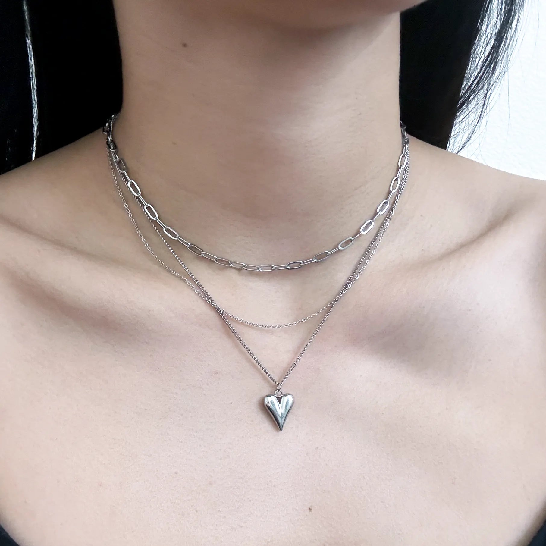 A close-up of a person's neck and upper chest wearing a layered silver necklace set, including a paperclip chain, a fine chain, and a pendant with a small heart-shaped cutout.
