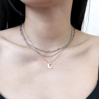 A close-up of a model's neck and upper chest wearing a layered silver necklace set. The set includes a thick paperclip chain, a fine chain, and a delicate chain with a small crescent moon pendant.