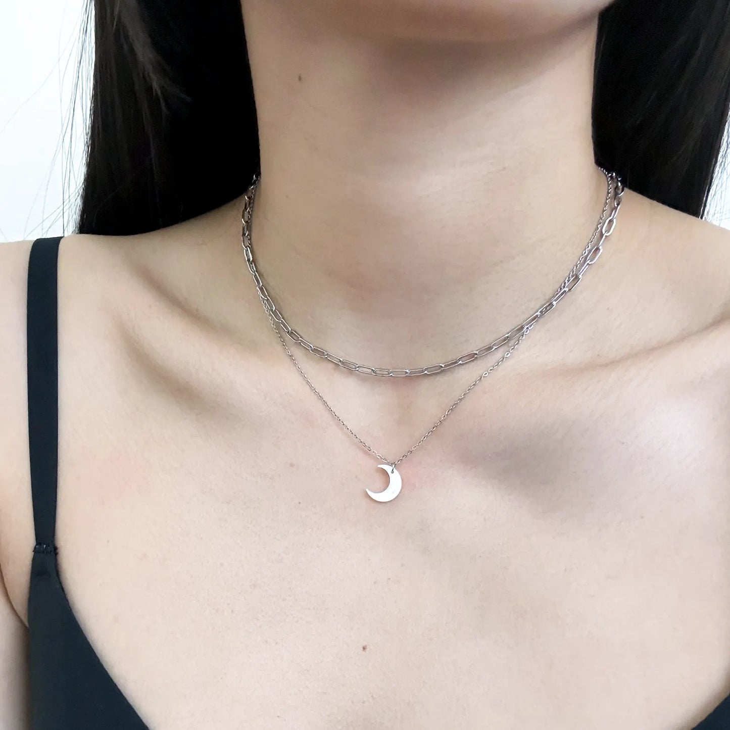 A close-up of a model's neck and chest wearing a layered silver necklace set, featuring a textured chain and a delicate chain with a small crescent moon pendant.