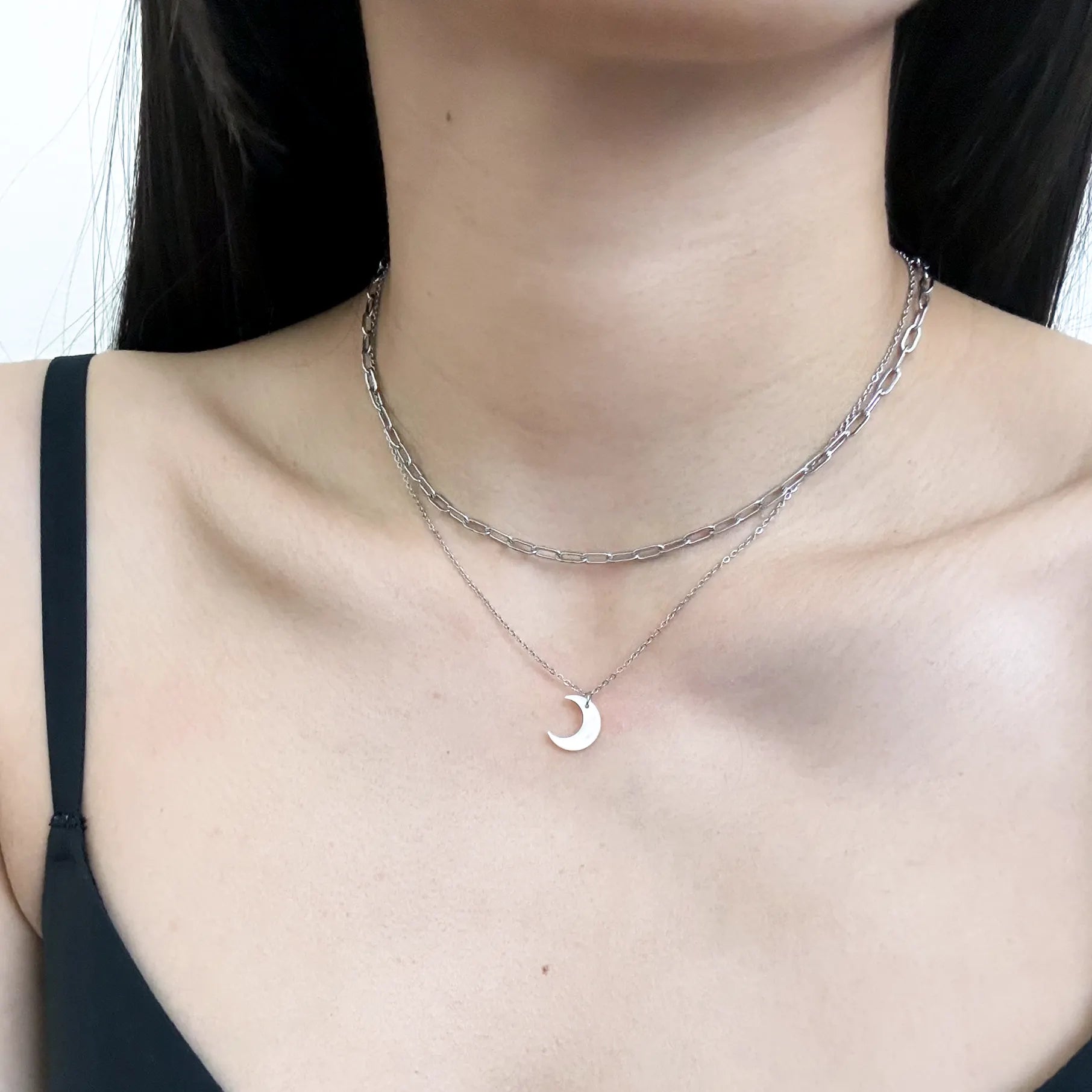 A close-up of a model's neck and chest wearing a layered silver necklace set, featuring a textured chain and a delicate chain with a small crescent moon pendant.