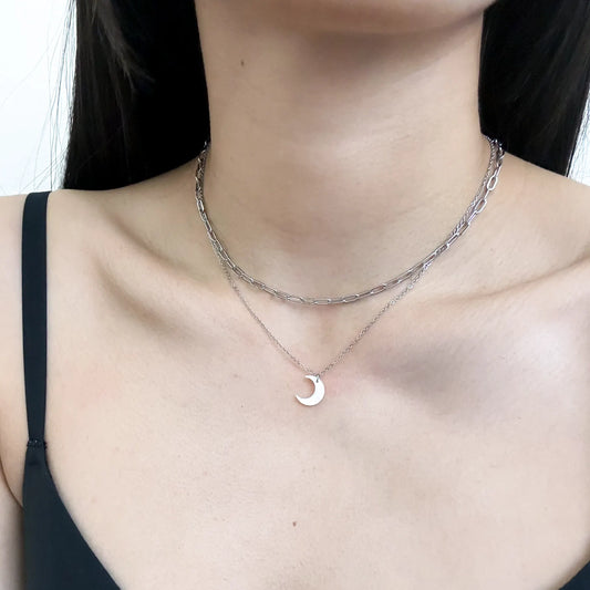 A close-up of a model's neck and chest wearing a layered silver necklace set, featuring a textured chain and a delicate chain with a small crescent moon pendant.