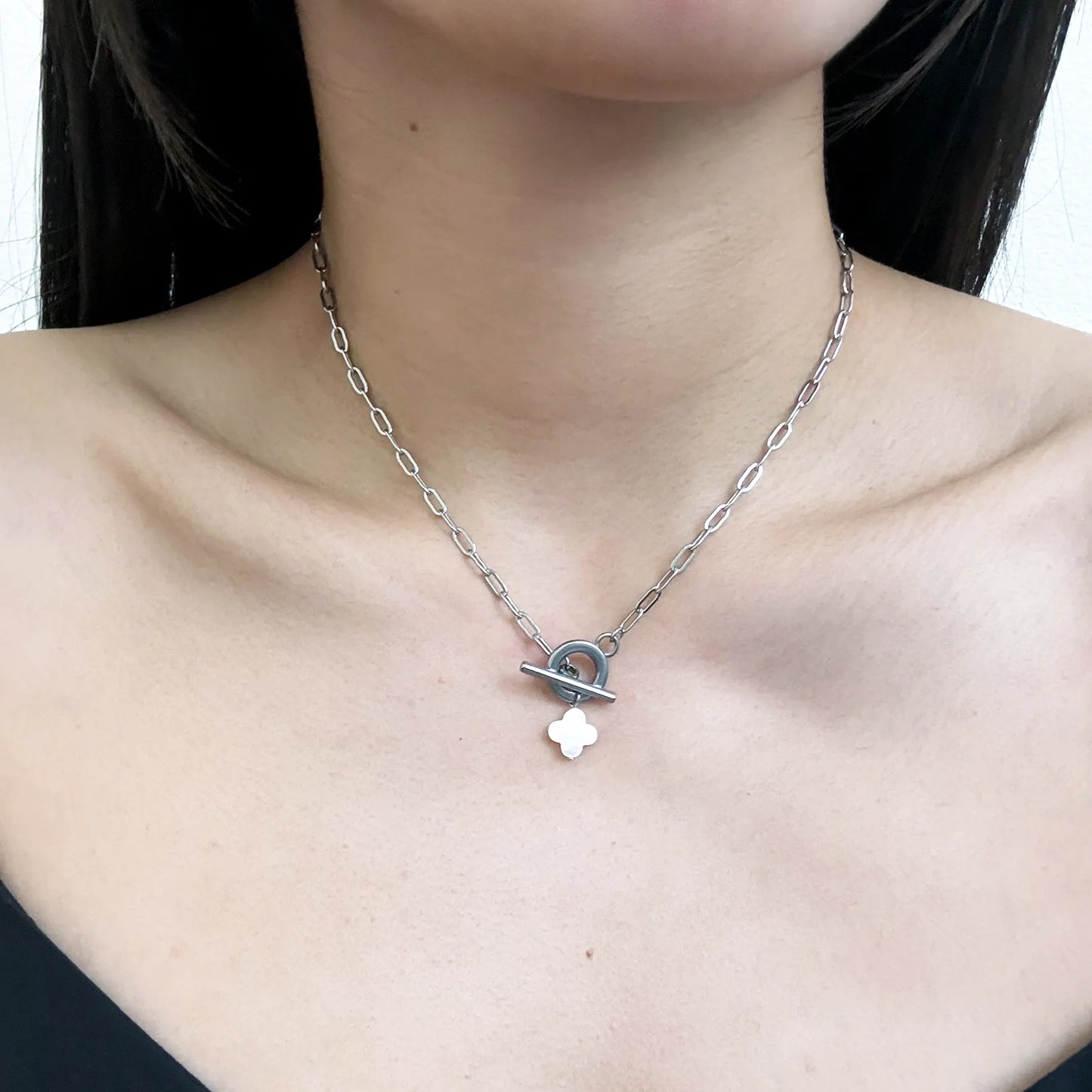 A close-up of a model's neck and chest wearing a silver chain necklace with a toggle clasp and a small white clover pendant.