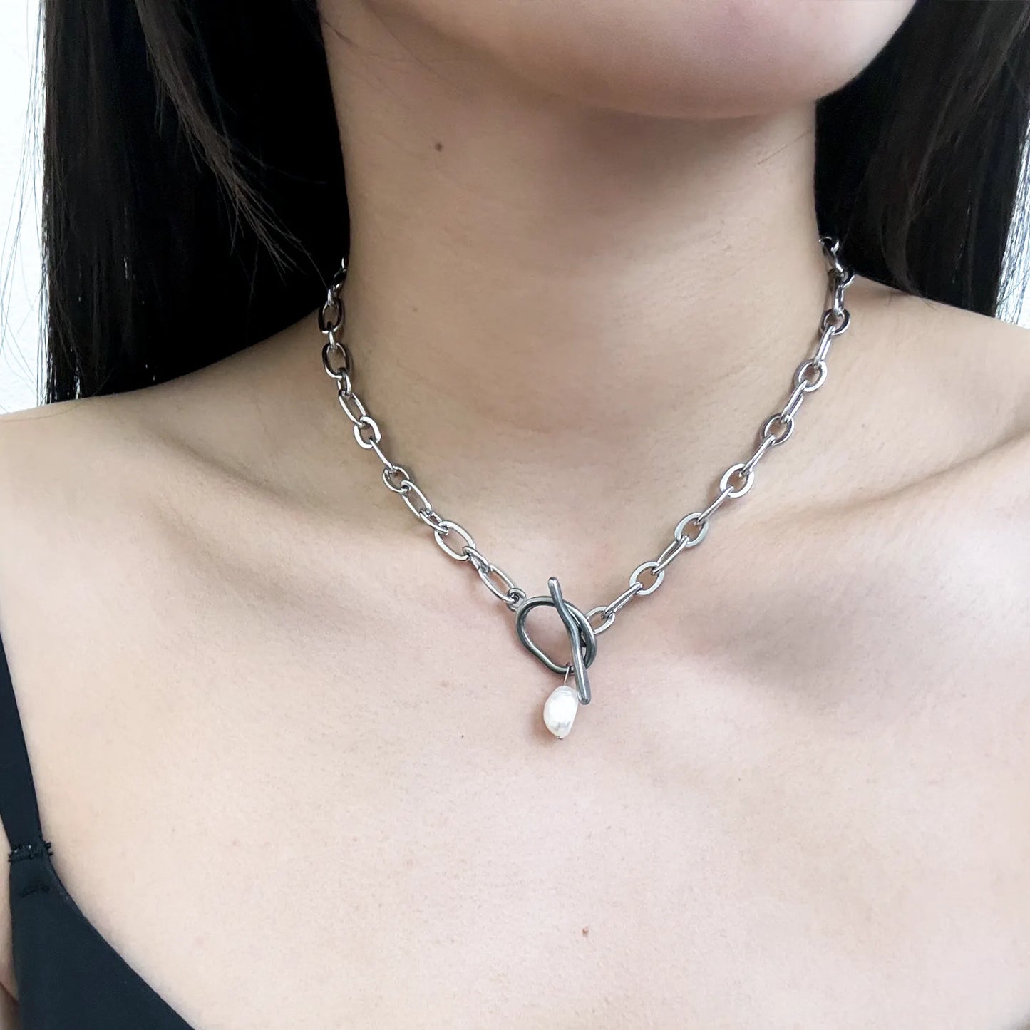 A close-up of a model's neck and chest wearing a silver chain necklace with a heart-shaped pendant and a small white teardrop charm.