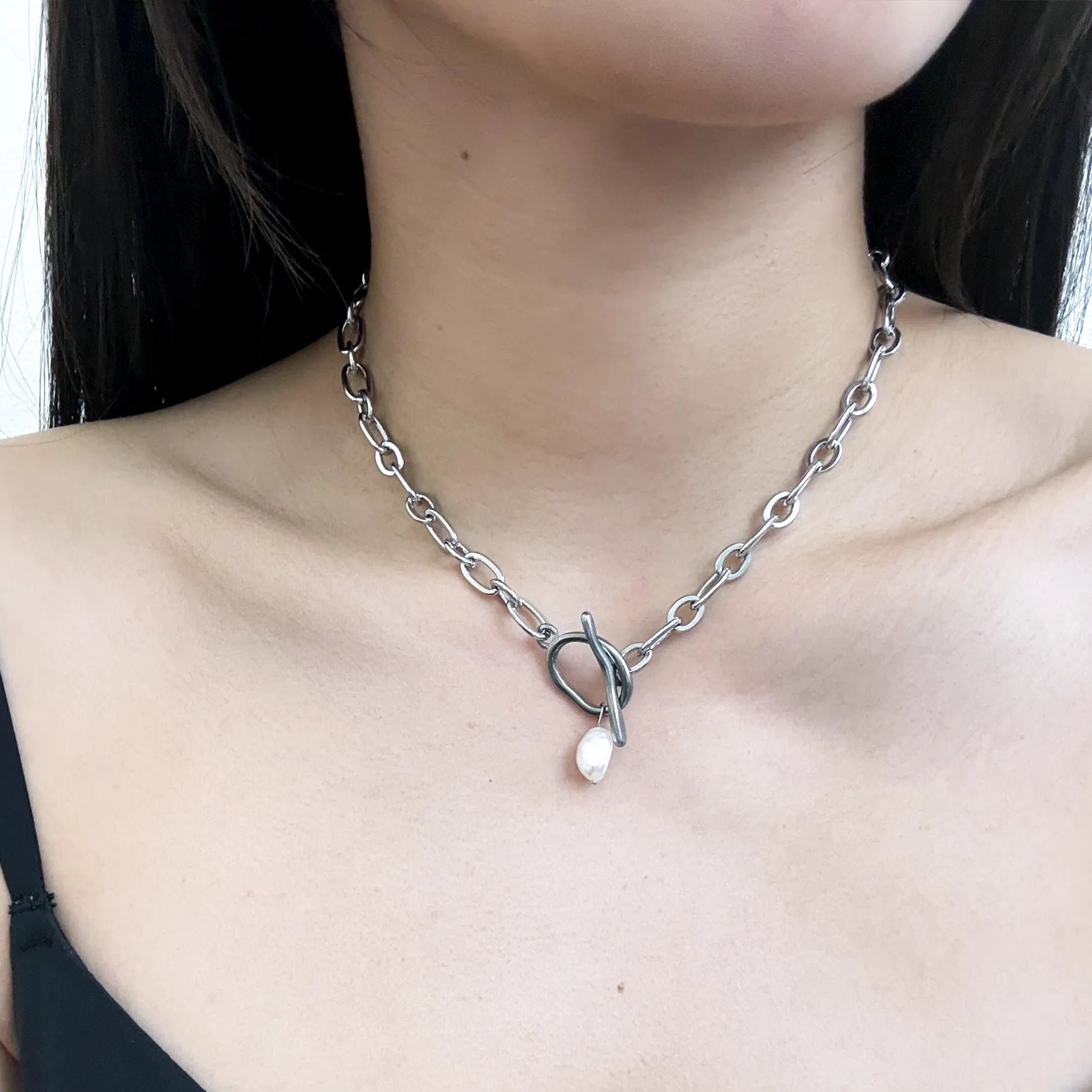 A close-up of a model's neck and chest wearing a silver chain necklace with a heart-shaped pendant and a small white teardrop charm.