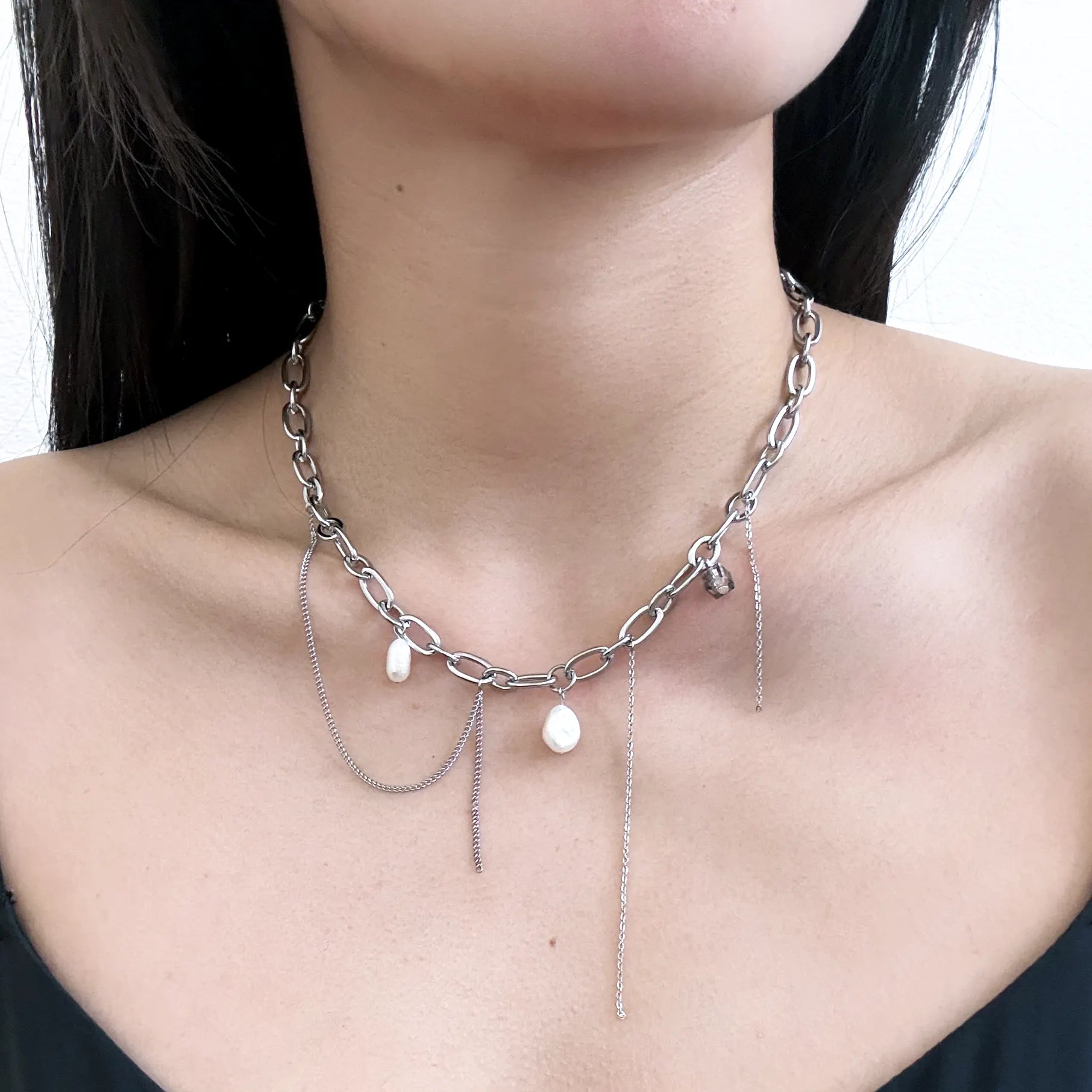 A close-up of a model's neck and chest wearing a silver-toned layered chain necklace with dangling pearl-like white beads and thin chain accents.